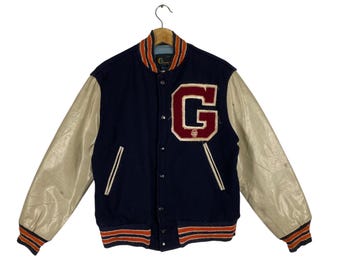 May include: A navy blue varsity jacket with cream-colored leather sleeves. The jacket features a large red and white "G" patch on the chest and orange and navy striped trim at the collar, cuffs, and waist. The jacket is hanging on a hanger.