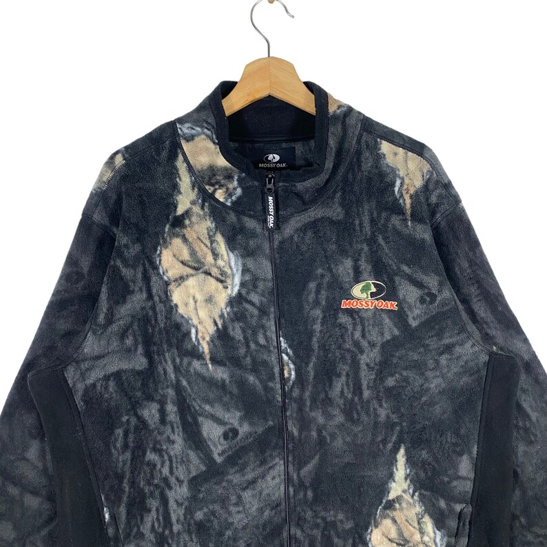 May include: A black fleece jacket with a camouflage pattern and the Mossy Oak logo. The jacket has a zipper closure and a stand-up collar.