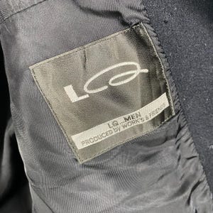 Pu&ograve; includere: Primo piano della fodera di una giacca blu navy scuro. Un'etichetta rettangolare con la scritta "Lq...MEN PRODUCED BY WORK'S & FRIENDS" &egrave; cucita nella fodera. L'etichetta &egrave; grigia con testo bianco e un logo bianco.