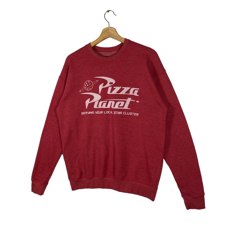 Puede incluir: Sudadera roja con un gr&aacute;fico blanco del logotipo de Pizza Planet de la pel&iacute;cula Toy Story. El logotipo dice "Pizza Planet" y "Serving your local star cluster".