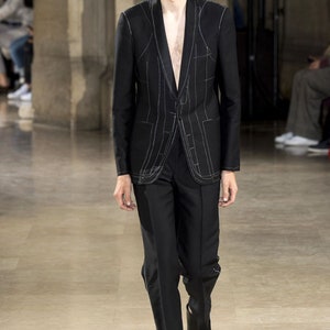May include: A black suit with white stitching detail. The jacket has a shawl collar and a single button closure. The trousers have a straight leg.