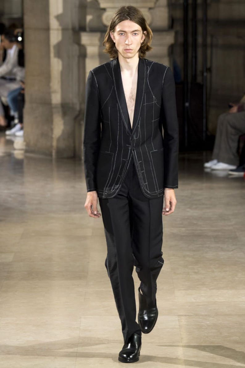 May include: A black suit with white stitching detail. The jacket has a shawl collar and a single button closure. The trousers have a straight leg.