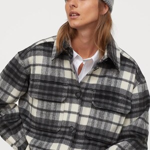 May include: A grey beanie and a black, white, and grey plaid shirt jacket with two front pockets and black buttons. The jacket is worn over a white collared shirt.