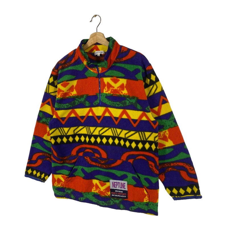 Puede incluir: Un jersey de forro polar colorido con un patr&oacute;n geom&eacute;trico en tonos de rojo, amarillo, verde, azul y negro. El jersey tiene una cremallera de media cremallera y un bolsillo canguro. El texto "NEPTUNE" est&aacute; impreso en el bolsillo.