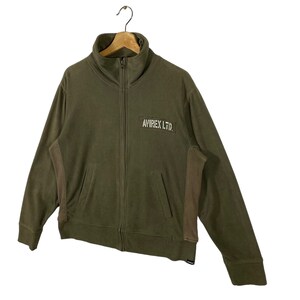 Puede incluir: Chaqueta con cremallera verde oliva con cuello alto y dos bolsillos delanteros. La chaqueta tiene un peque&ntilde;o parche rectangular en el pecho con el texto "AVIREX LTD." en blanco. La chaqueta est&aacute; colgada de una percha de madera.