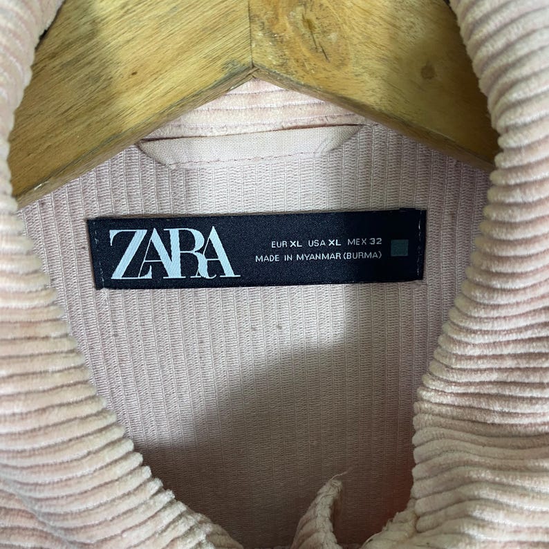 K&ouml;nnte beinhalten: Nahaufnahme einer hellrosa Cordjacke mit Kragen. Die Jacke hat ein Etikett mit der Aufschrift "ZARA" und Gr&ouml;&szlig;enangaben: EUR XL, USA XL, MEX 32. Hergestellt in Myanmar.