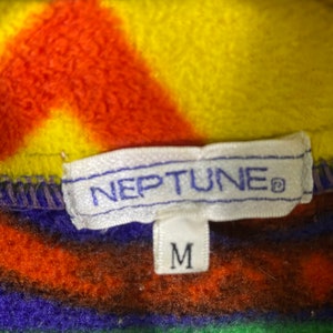 Puede incluir: Una etiqueta blanca con la palabra "NEPTUNE" en letras azules y un s&iacute;mbolo de marca registrada. La etiqueta est&aacute; unida a una tela de polar a rayas de colores con una etiqueta blanca m&aacute;s peque&ntilde;a que dice "M" en letras negras.