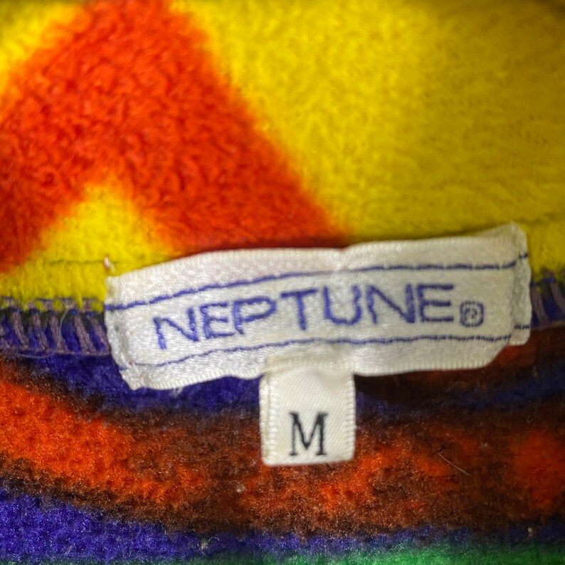 Puede incluir: Una etiqueta blanca con la palabra "NEPTUNE" en letras azules y un s&iacute;mbolo de marca registrada. La etiqueta est&aacute; unida a una tela de polar a rayas de colores con una etiqueta blanca m&aacute;s peque&ntilde;a que dice "M" en letras negras.
