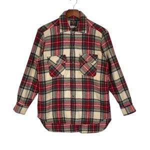 May include: A long-sleeve button-up shirt featuring a plaid pattern in red, cream, dark green, and black. The shirt includes a pointed collar, a front button placket, and two chest pockets with buttoned flaps, all displayed on a wooden hanger.