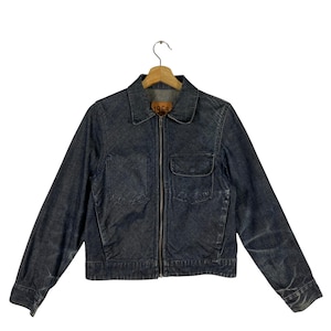 Puede incluir: Una chaqueta vaquera azul oscuro con cierre de cremallera y un pequeño bolsillo en el pecho izquierdo. La chaqueta tiene una etiqueta de cuero marrón con el texto "1969" en la parte delantera.