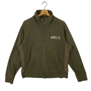 Puede incluir: Chaqueta con cremallera verde oliva con cuello alto y bolsillos laterales. La chaqueta presenta un parche en el pecho con el texto "AVIREX LTD." Las mangas y los laterales tienen un panel marr&oacute;n en contraste.