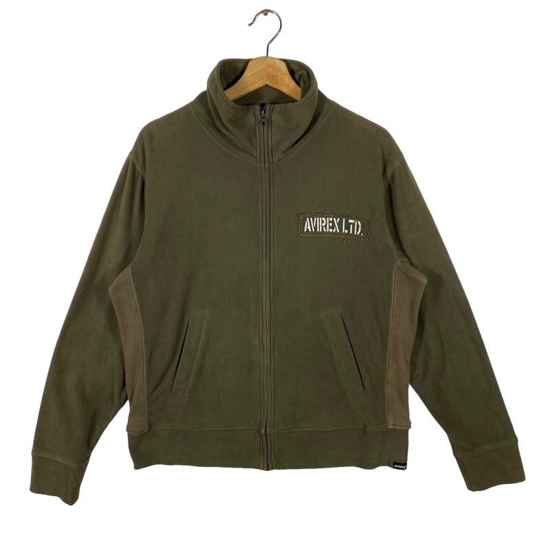 Puede incluir: Chaqueta con cremallera verde oliva con cuello alto y bolsillos laterales. La chaqueta presenta un parche en el pecho con el texto "AVIREX LTD." Las mangas y los laterales tienen un panel marr&oacute;n en contraste.