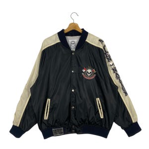 May include: Black and beige bomber jacket with a button-up front. The jacket features a logo patch on the chest and text on the sleeves that reads "ALIEN NATION". The jacket has a ribbed collar, cuffs, and hem.