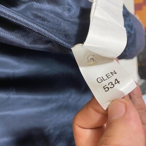 May include: Close-up of a navy blue fabric with a white tag. The tag reads "GLEN 534" in a sans-serif font. A small, round sticker with the word "contr&ocirc;le" is also visible. The fabric appears to be part of a garment.