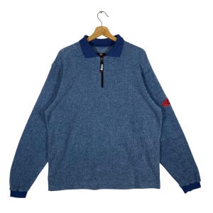 Può includere: Un pullover in pile blu con colletto e polsini blu scuro. La camicia a maniche lunghe ha una chiusura con zip a un quarto e una toppa con logo rosso sulla manica superiore sinistra. La camicia è appesa a una gruccia di legno.