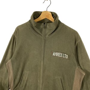 Puede incluir: Chaqueta polar verde oliva con cuello alto y cremallera completa. Cuenta con un parche marr&oacute;n con el texto "AVIREX LTD." en el pecho. La chaqueta tiene dos bolsillos delanteros y paneles marrones en los laterales.