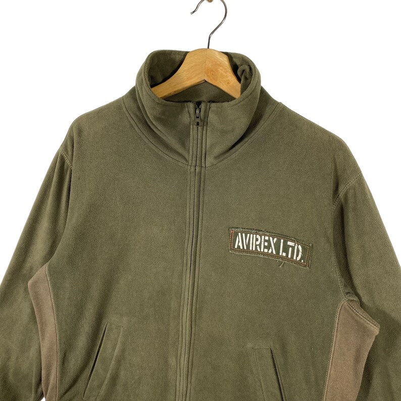 Puede incluir: Chaqueta polar verde oliva con cuello alto y cremallera completa. Cuenta con un parche marr&oacute;n con el texto "AVIREX LTD." en el pecho. La chaqueta tiene dos bolsillos delanteros y paneles marrones en los laterales.