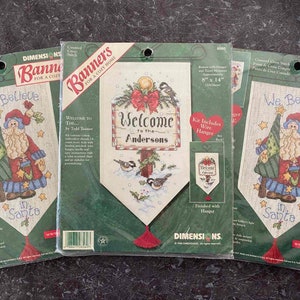May include: Three cross-stitch kits for banners featuring winter holiday themes. The kits include a wire hanger and are 8 inches by 14 inches. The designs include a Santa Claus, a welcome sign, and a winter scene with birds.