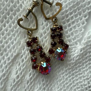 以下が含まれることがあります： ゴールドトーンの金属フレームが付いた、ヴィンテージのクリップオンイヤリング。各イヤリングは、小さな正方形の濃い赤色のラインストーンと、下部に大きな丸い虹色の赤いラインストーンが連なっています。イヤリングは、白いテクスチャード加工の背景にセットされています。