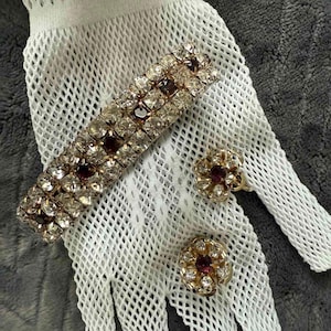 May include: A vintage jewelry set featuring a bracelet and two matching earrings. The bracelet and earrings have a gold-tone metal setting with clear and burgundy-colored rhinestones. The jewelry is displayed on a white, open-weave glove.
