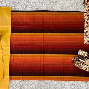 May include: A collection of fabric scraps in various shades of orange, red, and brown. The fabric is striped and has a gradient effect. There are also rolls of patterned wrapping paper with floral designs.