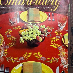 May include: Vintage embroidery book cover featuring a red tablecloth with yellow plates, silverware, and glassware. A bowl of fruit and a folded napkin are on the table. The book title "Embroidery" is in large gold letters.