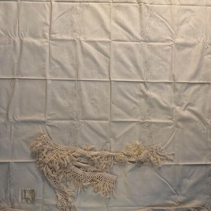 May include: Cream-colored fabric with a floral pattern and a decorative fringe trim. The fabric has a grid-like pattern and is adorned with delicate floral designs. The fringe trim adds a textural element to the textile.