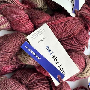 May include: Close-up of several skeins of Malabrigo Silky Merino yarn in shades of burgundy, brown, and pink. Each skein has a tag with the brand name and yarn details, including "173 STONECHAT". The yarn is hand-wash and dry flat.