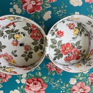 May include: Open, two-part vintage hatbox with a floral pattern of red, yellow, and pink roses on a white background. The box is open, revealing the interior design. The exterior is white with a silver trim. The box is on a floral patterned surface.