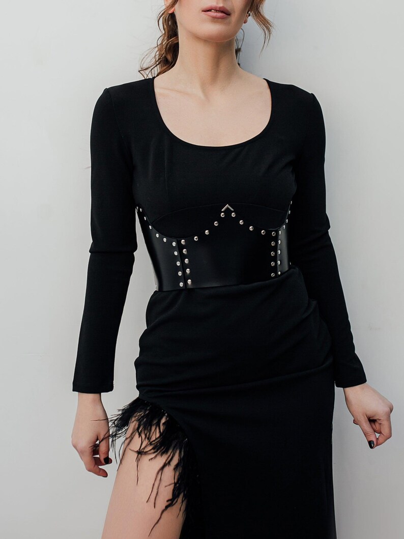 May include: A black long-sleeved dress with a scoop neckline and a high slit adorned with black feathers. A black leather corset with silver studs is worn over the dress, cinching the waist.