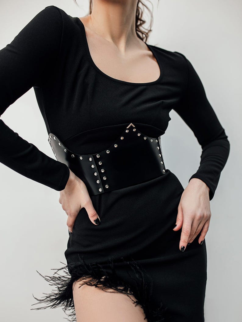 May include: A black corset belt with silver-colored studs is worn over a black long-sleeved dress. The dress has a scoop neckline and a feathered hemline. The belt is made of a smooth, dark material.