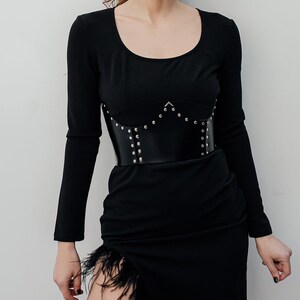 May include: A black long-sleeved dress with a scoop neckline and a high slit adorned with feathers. A black leather corset-style belt with silver studs cinches the waist, creating a defined silhouette. The dress is a solid color.
