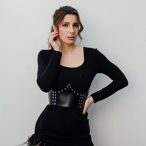 May include: A black dress with long sleeves and a square neckline. The dress features a wide, black leather belt with silver studs. The skirt has a feathered trim along the hemline. The woman is touching her hair.