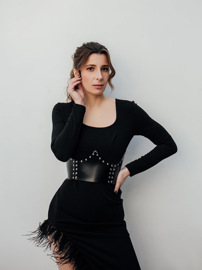 May include: A black dress with long sleeves and a square neckline. The dress features a wide, black leather belt with silver studs. The skirt has a feathered trim along the hemline. The woman is touching her hair.
