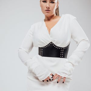 May include: A woman wearing a white long-sleeved dress with a black studded corset. The corset cinches the waist, creating a defined silhouette. The dress has a V-neckline and fitted sleeves. The woman has blonde hair pulled back.