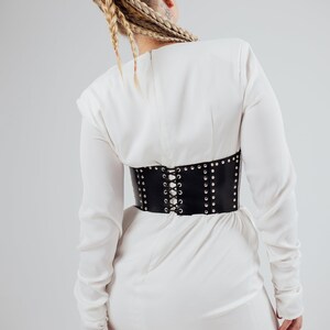 May include: A black leather corset belt with silver-colored studs and a lace-up back is worn over a white dress. The corset cinches the waist, creating a defined silhouette. The dress has long sleeves and a simple design.