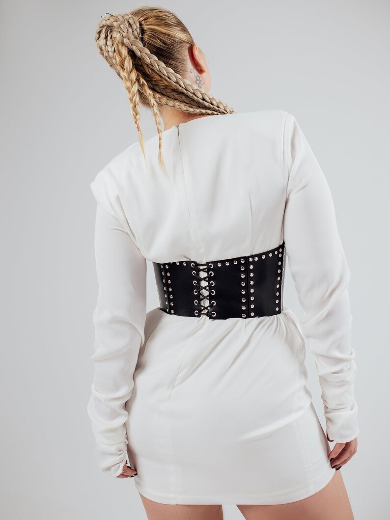 May include: A black leather corset belt with silver-colored studs and a lace-up back is worn over a white dress. The corset cinches the waist, creating a defined silhouette. The dress has long sleeves and a simple design.