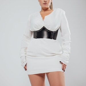 May include: A white long-sleeved dress with a V-neckline and a black leather corset. The corset is adorned with silver studs. The dress has a fitted silhouette and long sleeves.