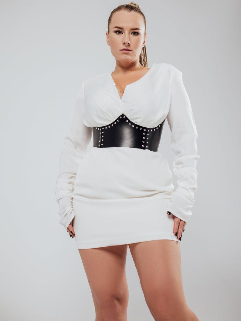 May include: A white long-sleeved dress with a V-neckline and a black leather corset. The corset is adorned with silver studs. The dress has a fitted silhouette and long sleeves.