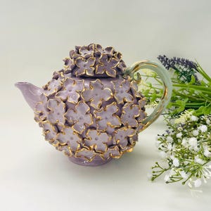 Puede incluir: Una tetera morada con un diseño floral y detalles dorados. La tetera está hecha de cerámica y tiene un aspecto delicado y femenino.