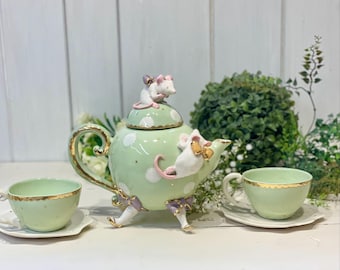 Pode incluir: Uma chaleira verde e branca com um padrão de bolinhas e uma figura de rato branco no topo. A chaleira tem detalhes dourados e está sentada em uma mesa com duas xícaras de chá e pires combinando.