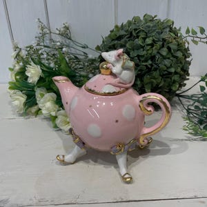 Könnte beinhalten: Eine rosafarbene Teekanne mit weißen Punkten und goldenen Verzierungen. Die Teekanne hat drei Beine und ist mit violetten Bändern verziert. Eine kleine weiße Maus mit einem goldenen Paket sitzt auf dem Deckel der Teekanne.