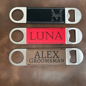 May include: Three personalized bottle openers with leatherette handles. The top opener is black with the name "Murray" and an initial "M". The middle opener is red with the name "LUNA". The bottom opener is brown with the name "ALEX GROOMSMAN".