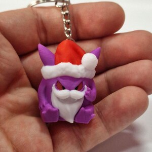 Può includere: Portachiavi natalizio viola con un cappello di Babbo Natale bianco e una barba bianca. Il portachiavi ha la forma di un personaggio dei cartoni animati con le corna e gli occhi arrabbiati.