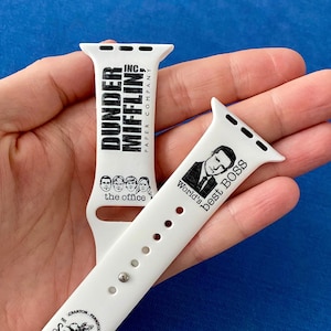 May include: Two white Apple Watch bands with black text and images. The top band says "Dunder Mifflin Inc. Paper Company" and the bottom band says "World's Best Boss" with a picture of Michael Scott. The bottom band also says "Schrute Farms" with a small image of a beet.