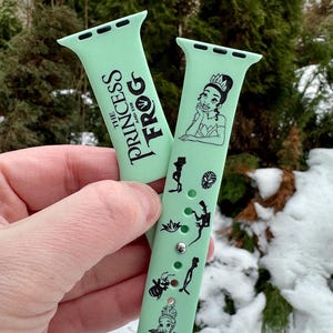 May include: Two pale green watch straps with black graphics and text. One strap has the text "THE PRINCESS AND THE FROG". The other strap features a princess illustration and other character graphics. The straps are held in front of a snowy background.