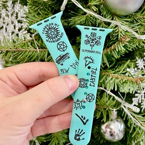 May include: Two turquoise blue Apple Watch bands with black designs. One band has the text "Carry On My Wayward Son" and the other has the text "Supernatural" and "Castiel".