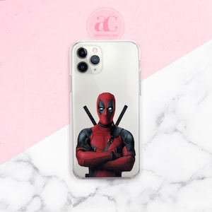 Puede incluir: Una funda transparente para teléfono con una imagen impresa de Deadpool, un personaje de superhéroe, con los brazos cruzados. La funda está diseñada para un iPhone 11 Pro.