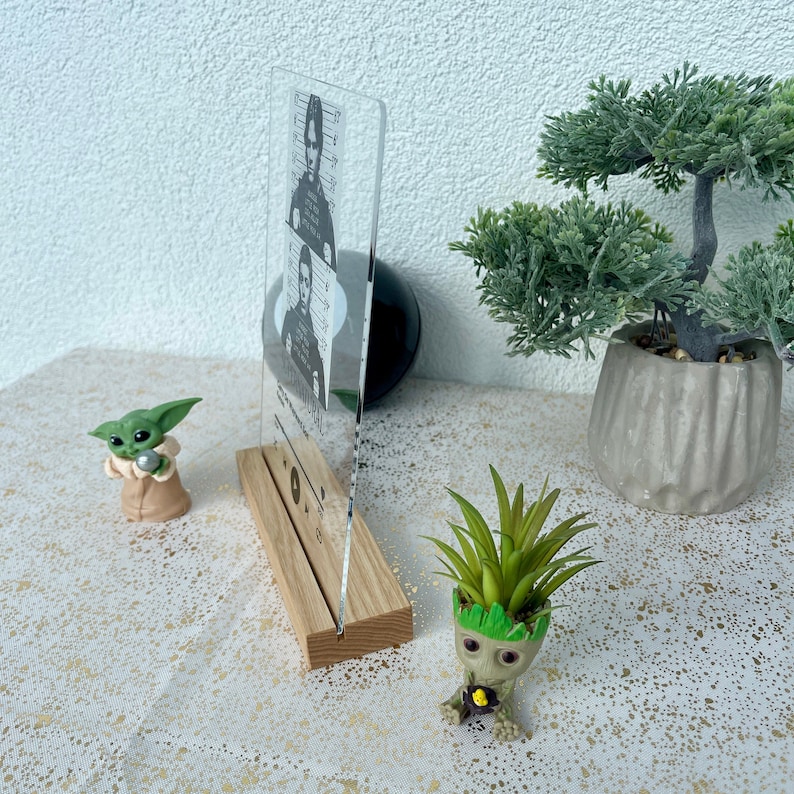 May include: A clear acrylic photo frame with a wooden base. The frame features a black and white photo of a man with a moustache. The frame is sitting on a white surface with a small green plant in a pot.