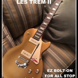 May include: A gold-colored electric guitar with a cream-colored pickguard and pickups. The guitar has a silver tremolo system and tuning pegs. Text on the image reads "DUESENBERG/GOLDO LES TREM II" and "EZ BOLT-ON FOR ALL STOP TAIL GUITARS."